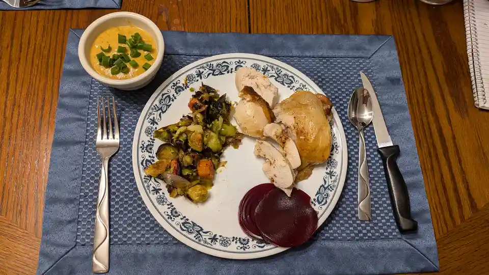 A top-down photo of a plated meal on a blue placemat, featuring sliced roasted chicken, roasted brussels sprouts and…