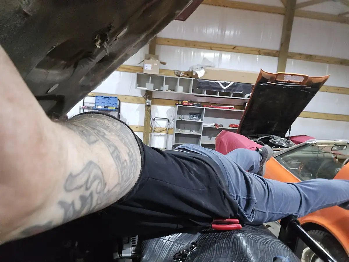 A person with a tattooed arm is lying on their back under a lifted car in a garage, working on the underside. The car's hood…