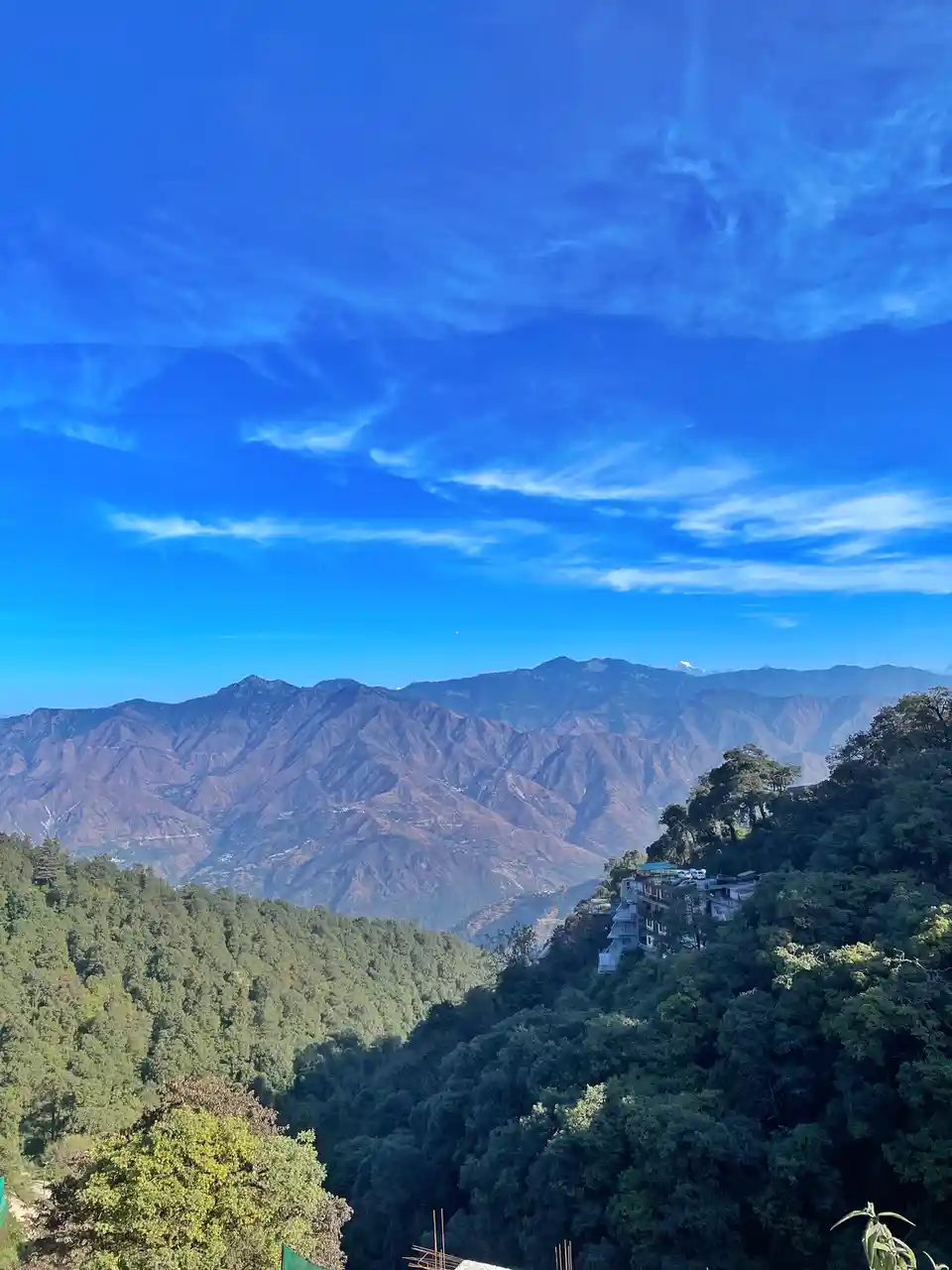 A scenic mountain landscape with lush green forests, layered mountain ranges under a vibrant blue sky with wispy clouds, and…