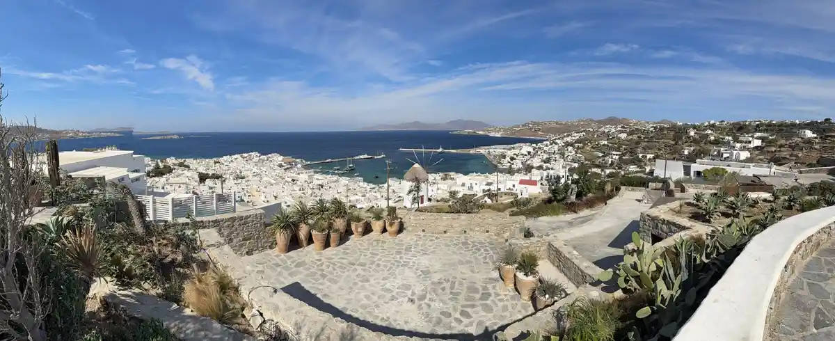 A panoramic photo of a sunny, scenic Greek island town with white buildings, a blue sea, and a stone patio with potted…