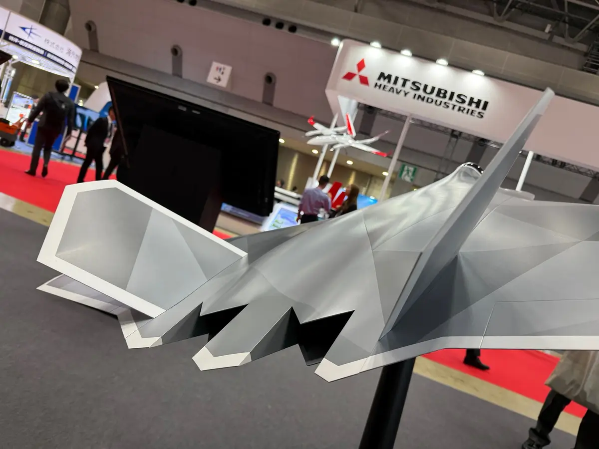 a low-angle shot of a stylized, angular gray fighter jet model on display at a Mitsubishi Heavy Industries booth, with…