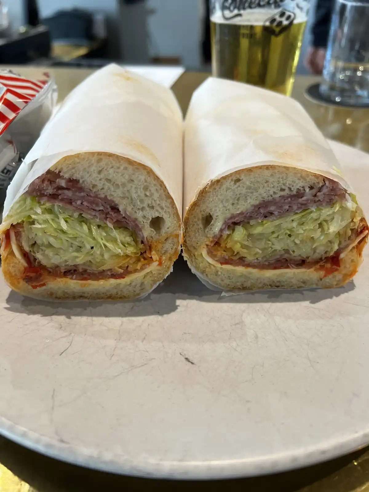 A close-up photo shows a sandwich cut in half, revealing layers of meat, lettuce, and sauce inside a crusty roll, resting on…
