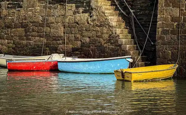 Several small, colorful rowboats — red, blue, and yellow — are moored in calm water beside a weathered stone wall with a…