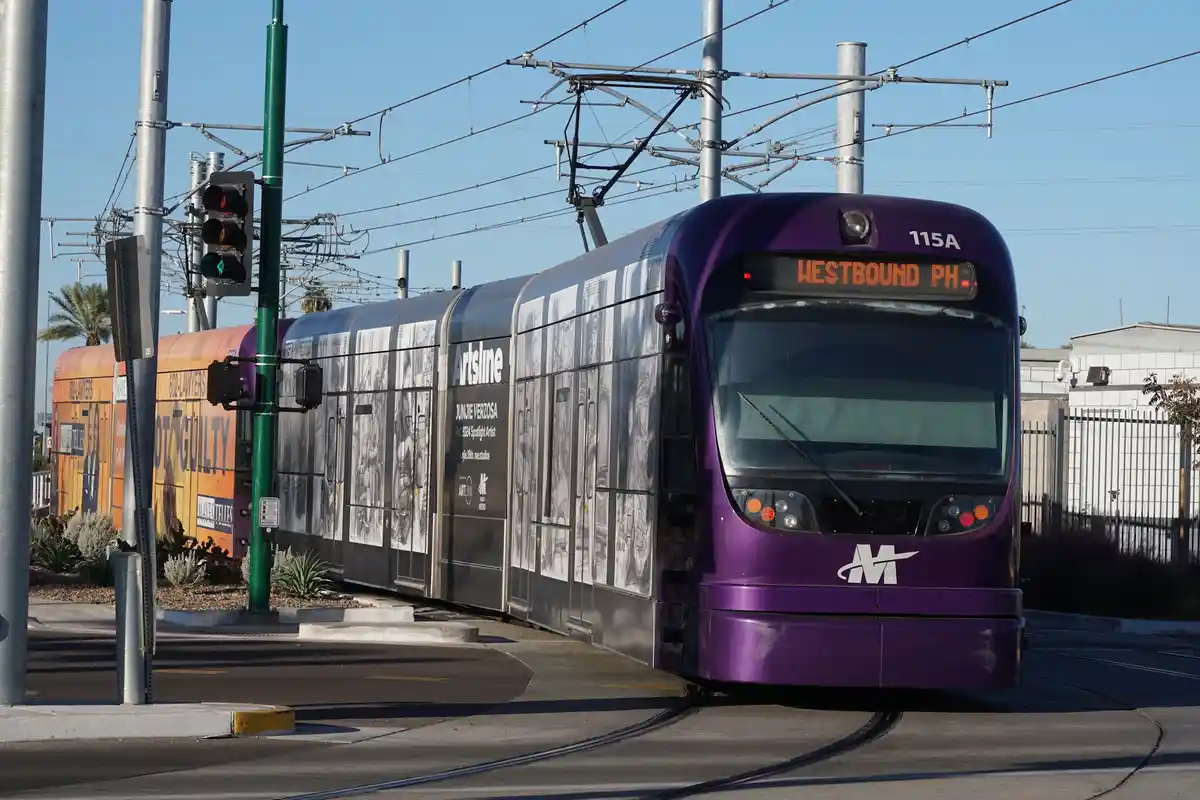 A purple and silver light rail train, numbered 115A, is shown on tracks under a clear blue sky. The front display reads…