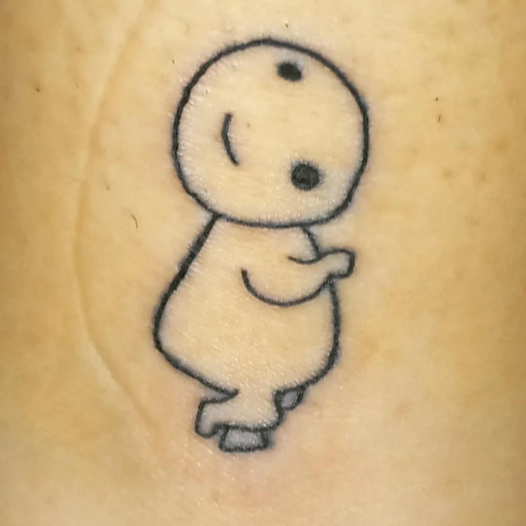 A close-up photo shows a simple black ink tattoo on light skin, depicting a stylized baby with a round head, one eye, a…