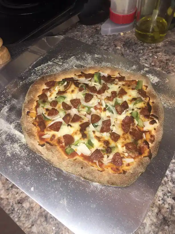 A homemade pizza with pepperoni, green peppers, and onions sits on a floured metal pizza peel on a granite countertop, with…