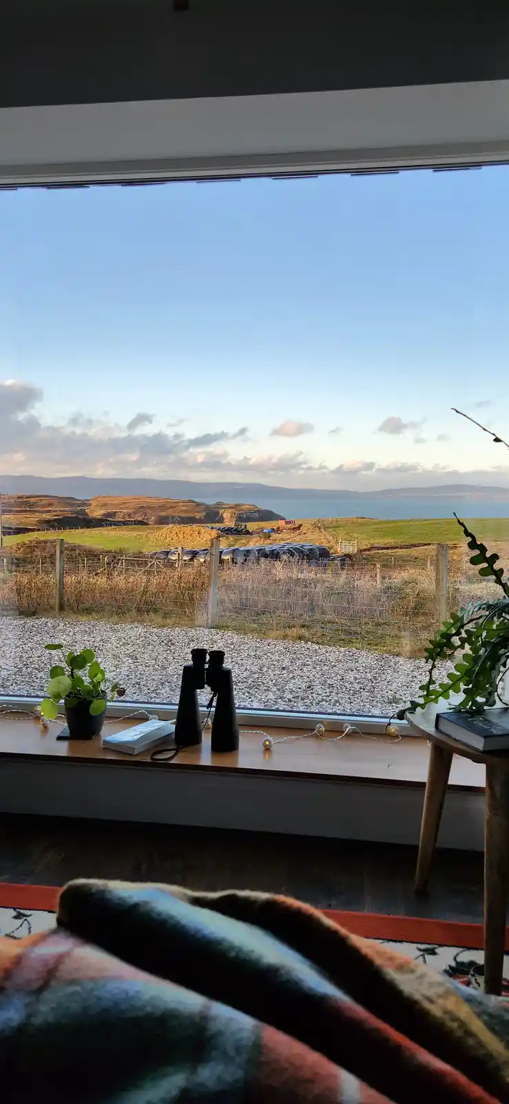 A cozy indoor scene looking out a large window at a scenic coastal landscape with hills, sea, and a gravel path. On the…
