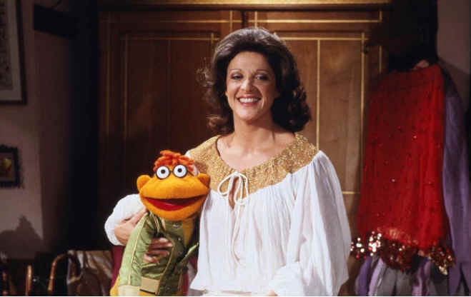 a woman with dark curly hair and a white blouse with gold trim smiles while holding a puppet with an orange face, large…