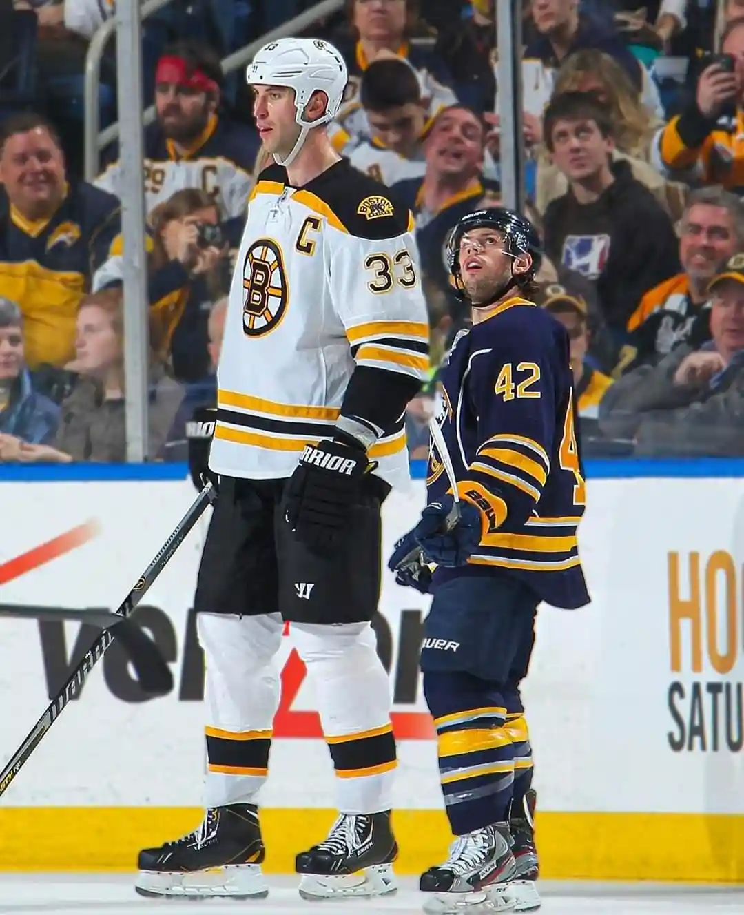 Two hockey players stand on the ice; the taller one in a white Boston Bruins jersey with 'C' and number '33' looks forward…