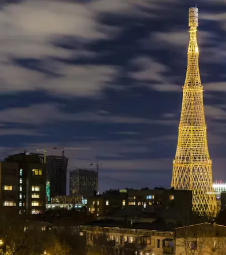 A nighttime cityscape photo shows a tall, illuminated lattice tower dominating the skyline, with other buildings and cranes…
