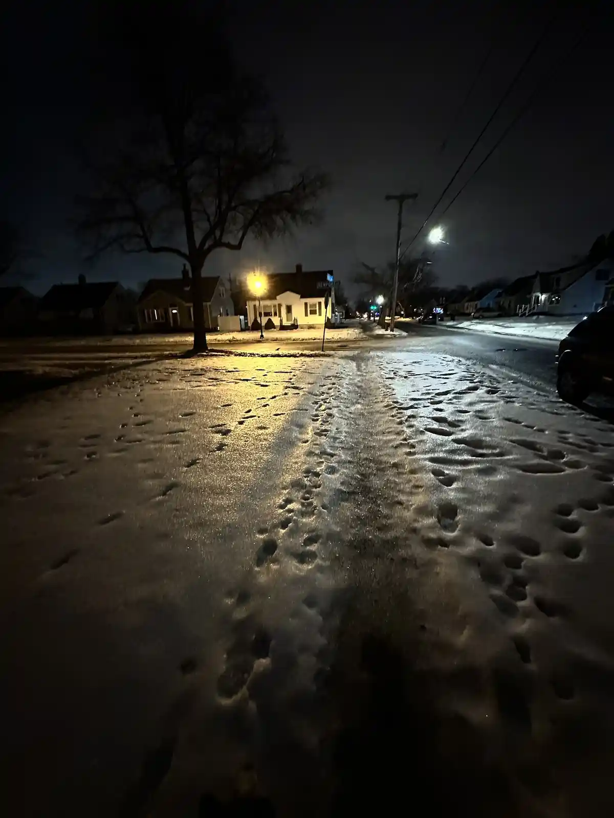 A nighttime photo of a snow-covered residential street with footprints leading toward the camera, illuminated by…