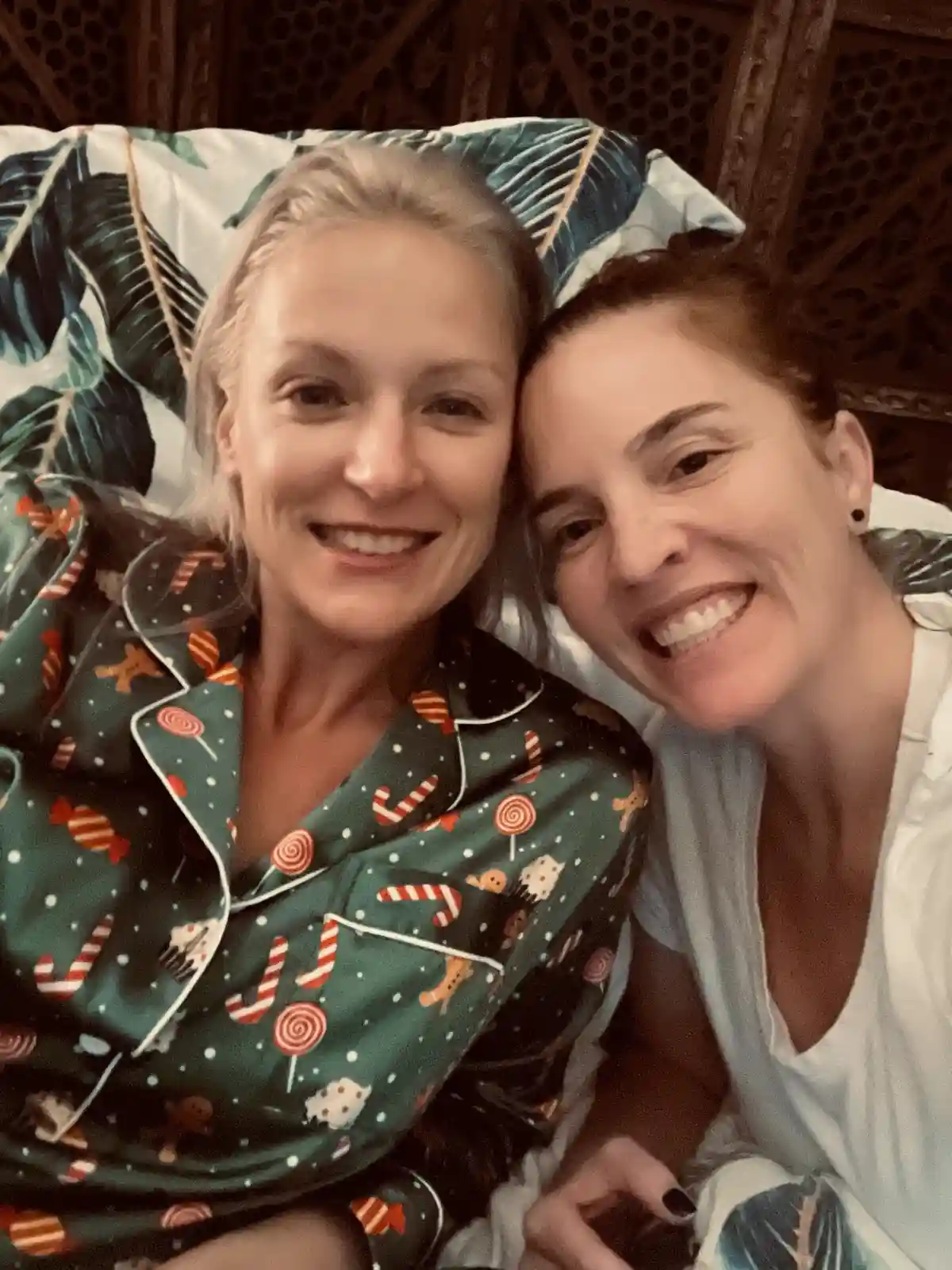 Two smiling women are lying close together on a bed with patterned pillows, one wearing festive holiday-themed pajamas and…