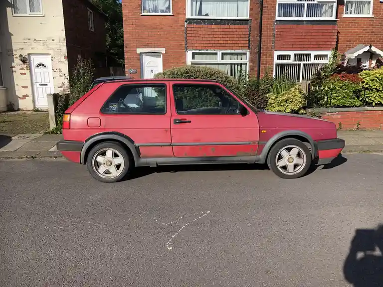 A red Volkswagen Golf GTI is parked on a residential street, showing visible rust and wear on its body panels, with a '106.1…