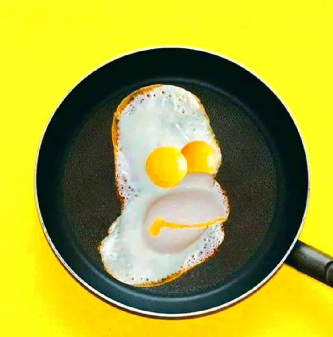 A fried egg in a black frying pan is shaped and colored to resemble the cartoon character Homer Simpson, with two yellow…