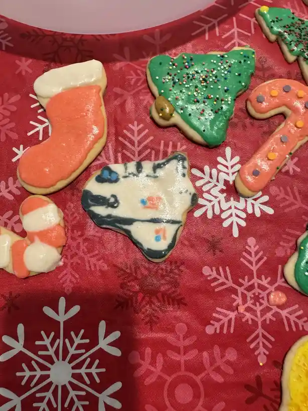 A close-up photo shows several Christmas-themed sugar cookies on a red fabric with white and pink snowflake patterns. The…