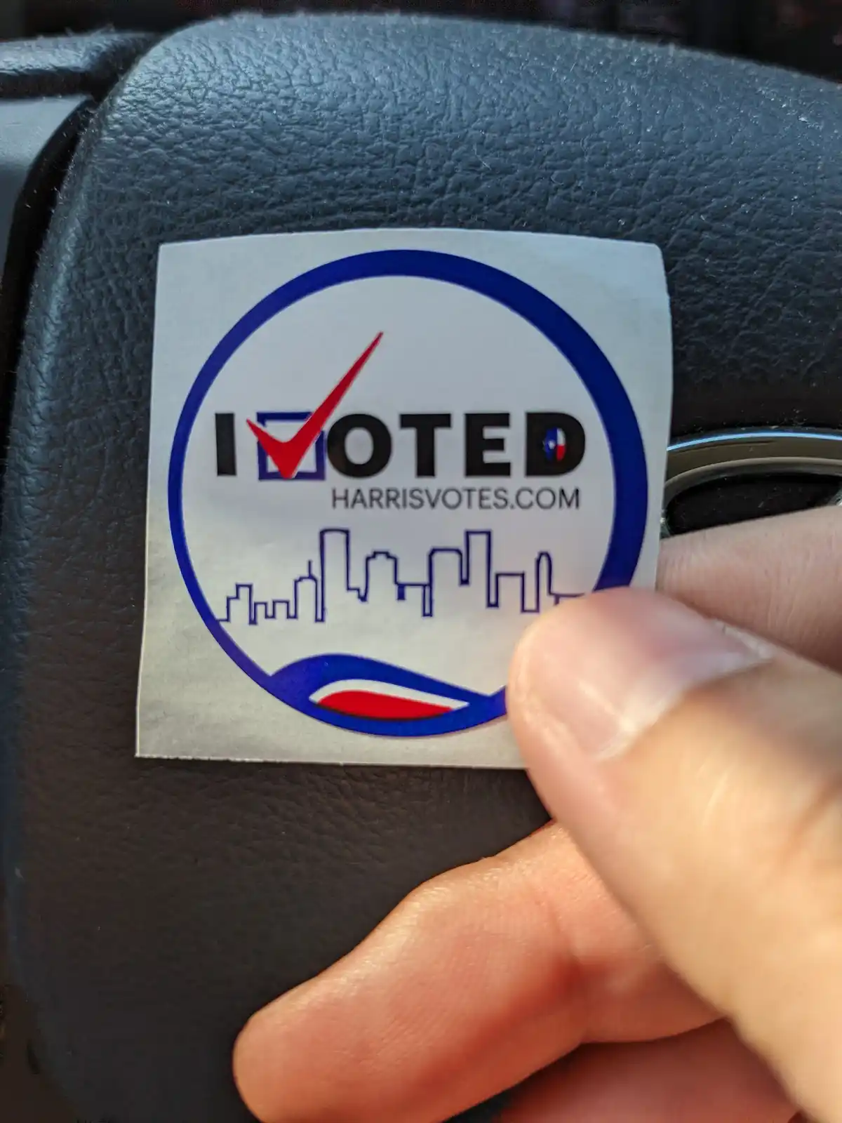 A close-up photo shows a hand holding an 'I VOTED' sticker from Harris County, Texas, which features a red checkmark over…