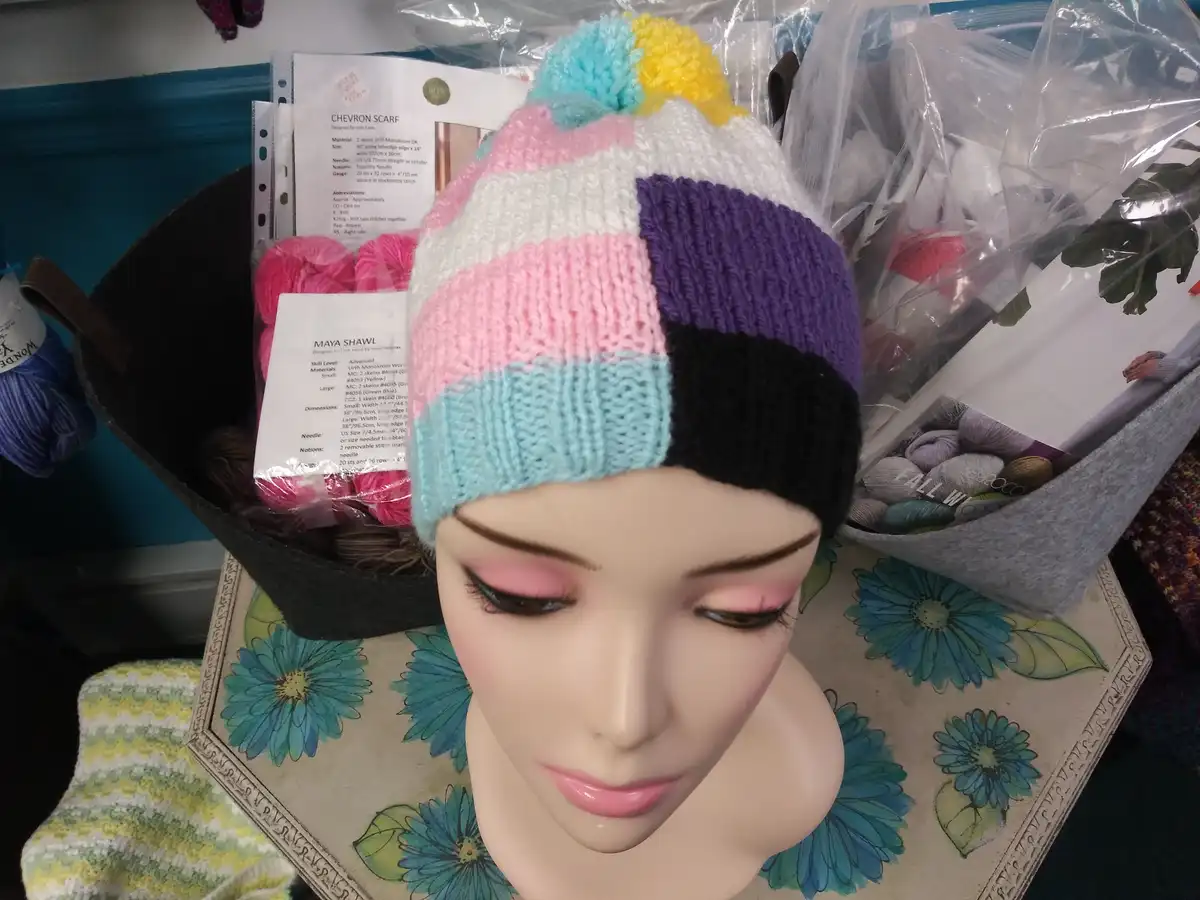 A mannequin head wearing a colorful, hand-knitted beanie with a rainbow of stripes and a multi-colored pom-pom sits on a…