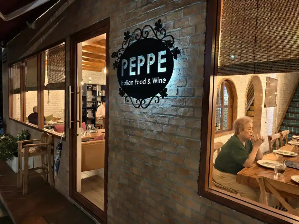 A photo of an Italian restaurant named 'PEPPE Italian Food & Wine' with a black circular sign on a brick wall. Inside, an…
