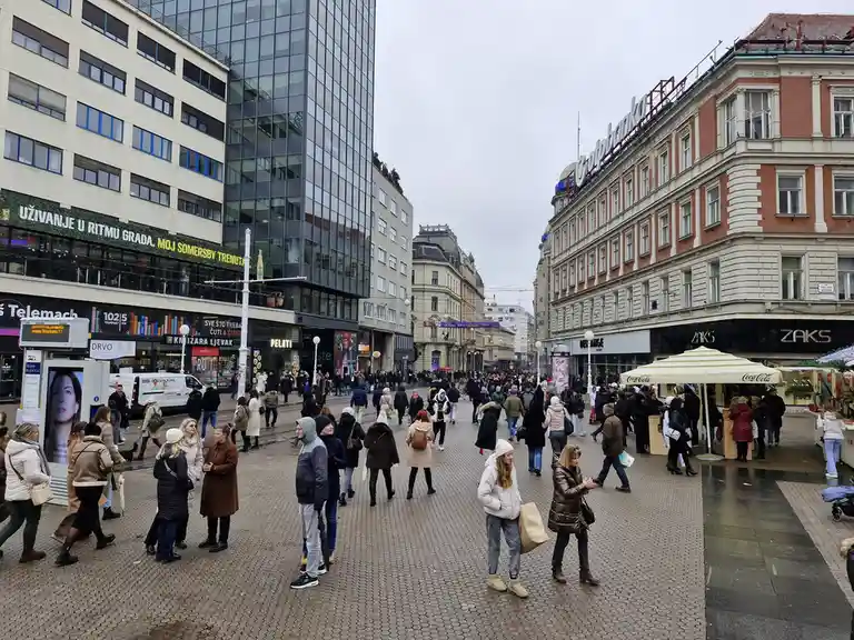 A crowded pedestrian street with many people walking or standing, flanked by modern and older buildings with visible…