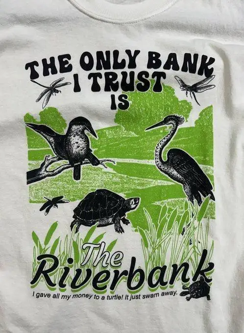 A white t-shirt graphic shows black-and-white illustrations of birds, a turtle, and dragonflies against a green riverbank…
