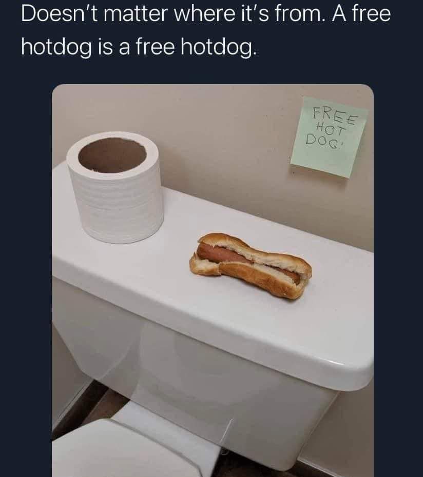 A hot dog sits on the lid of a white toilet tank next to an empty toilet paper roll. A green sticky note on the wall behind…