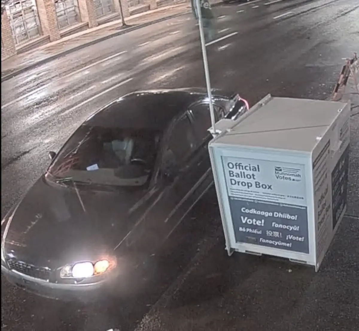 A dark car is parked next to an official ballot drop box on a wet street at night. The box has multilingual text including…