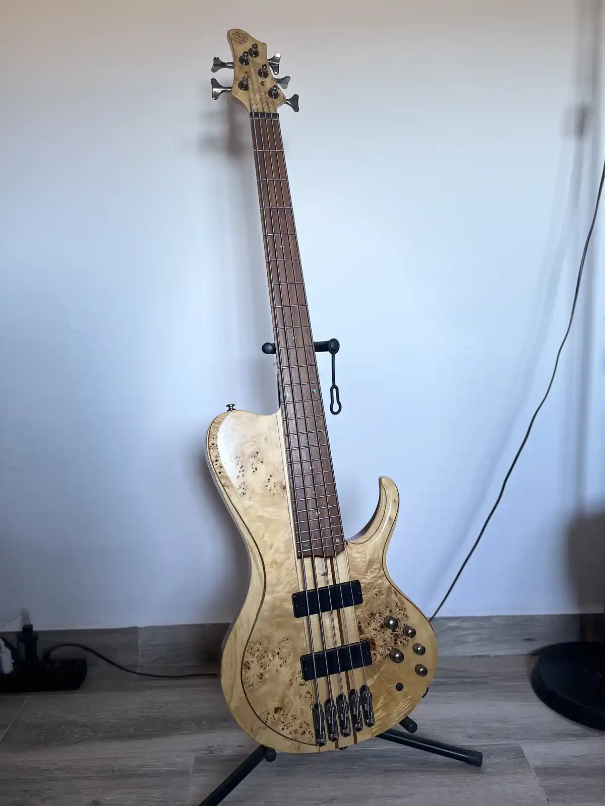 A light-colored, burl-finished 5-string electric bass guitar with a dark fretboard and chrome hardware stands upright on a…