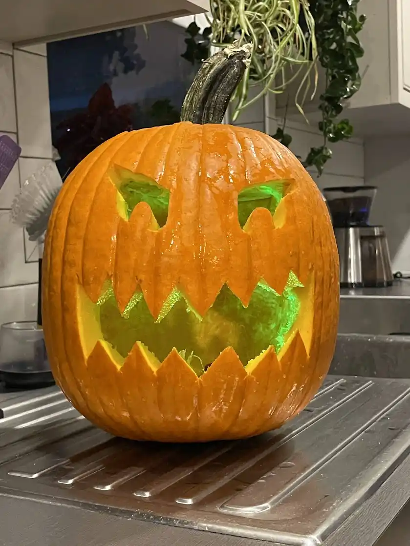 A close-up photo of a carved Halloween pumpkin sitting on a kitchen counter, illuminated from within by a bright green light…