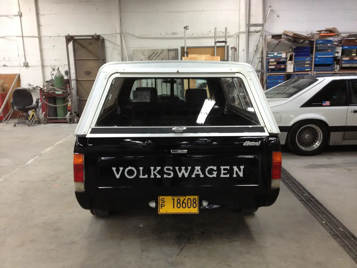 The rear view of a black and white vintage Volkswagen pickup truck parked in a garage, with the word 'VOLKSWAGEN'…