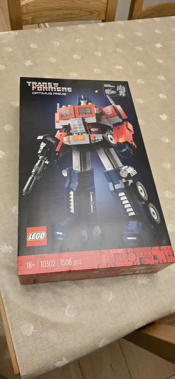 A photo of a LEGO Transformers Optimus Prime set box (set #10302, 1508 pieces, 18+) resting on a patterned tablecloth. The…
