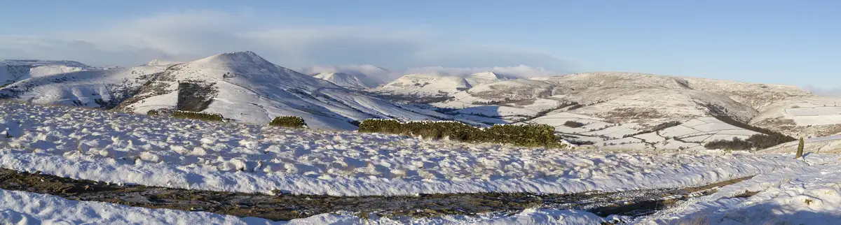 A wide, snowy landscape featuring rolling hills covered in white snow under a clear blue sky, with stone walls and patches…
