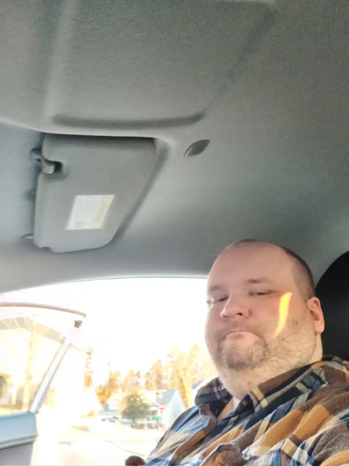 A man with a short beard and plaid shirt is sitting in a car, looking drowsy with his eyes half-closed. Sunlight streams in…