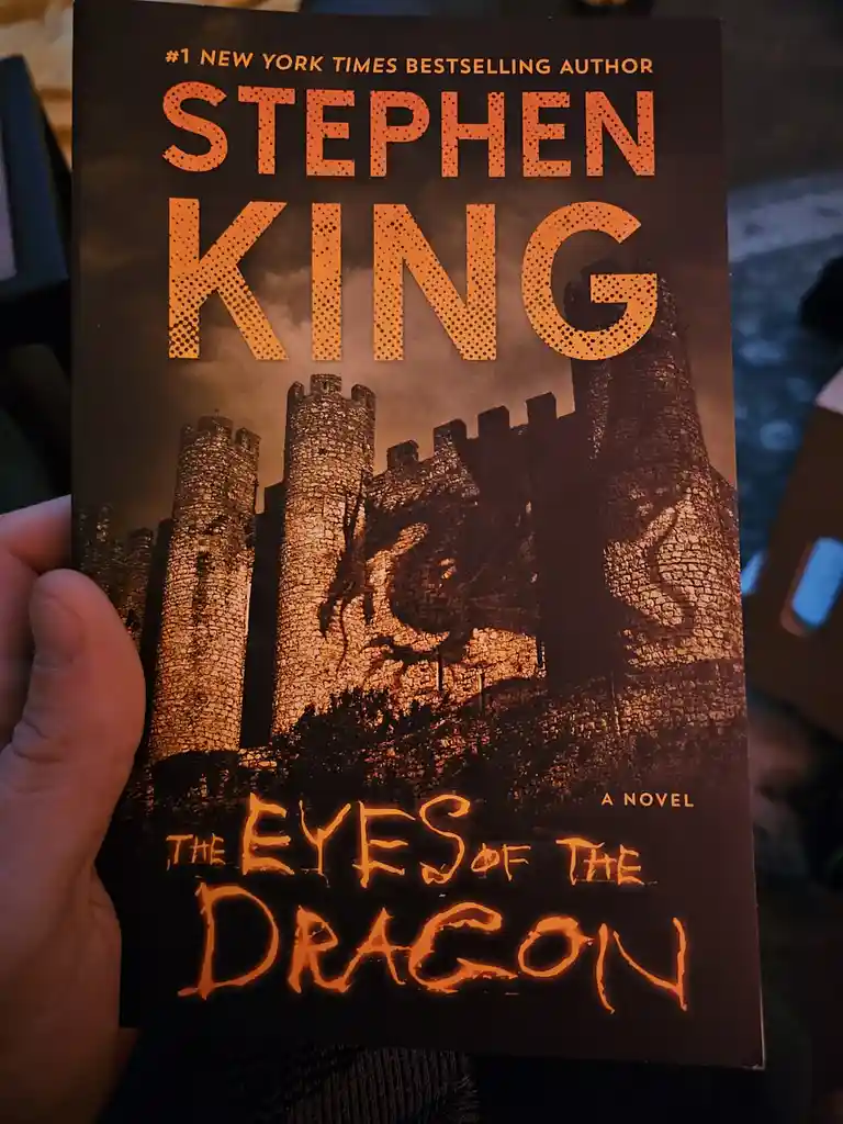 A hand holds a Stephen King novel titled 'The Eyes of the Dragon,' featuring a dark, orange-toned cover with a castle and a…