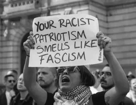 A black-and-white photo shows a person at a protest holding a handmade sign that reads 'YOUR RACIST PATRIOTISM SMELLS LIKE…