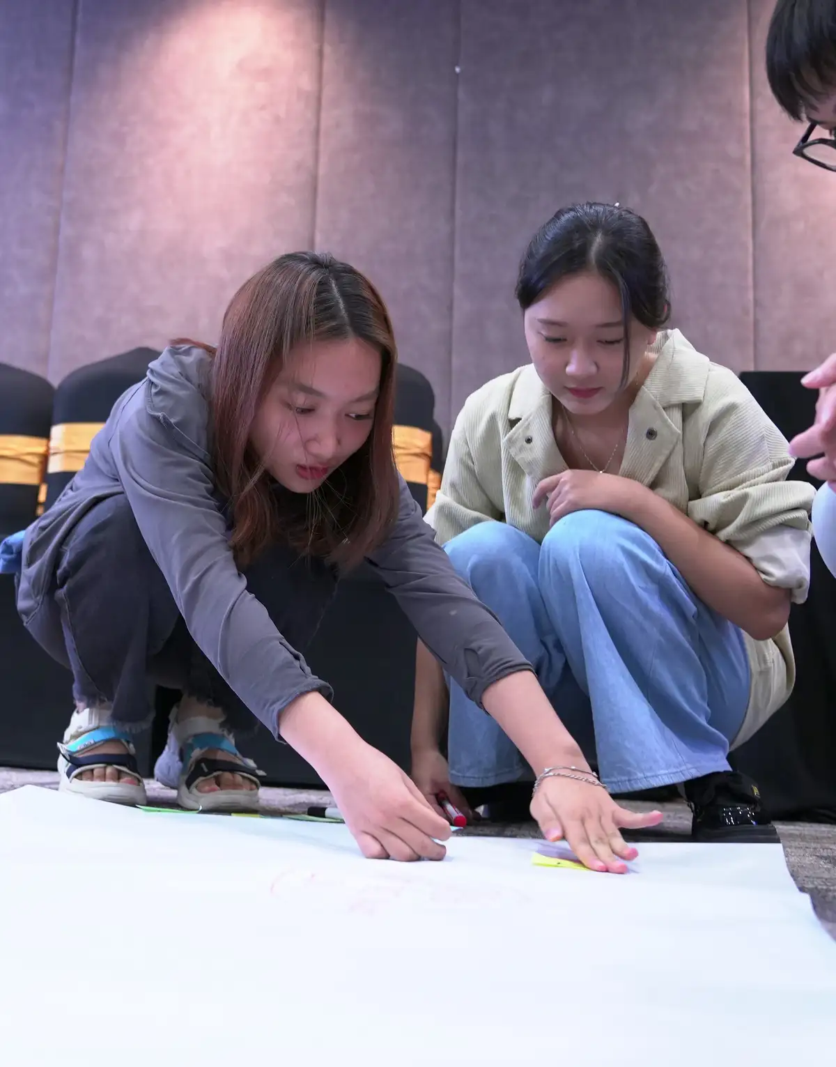 Two young women are crouched on the floor, intently focused on drawing or writing on a large white sheet of paper. One woman…
