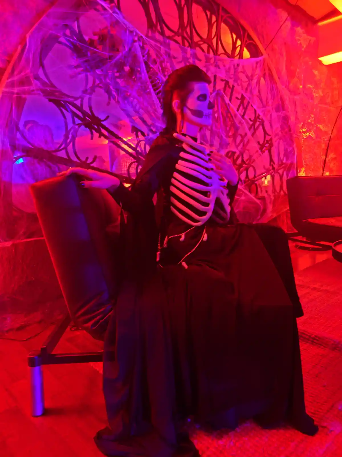 A person dressed in a skeleton costume with white ribcage and skull makeup sits elegantly on a black chair in a dimly lit…