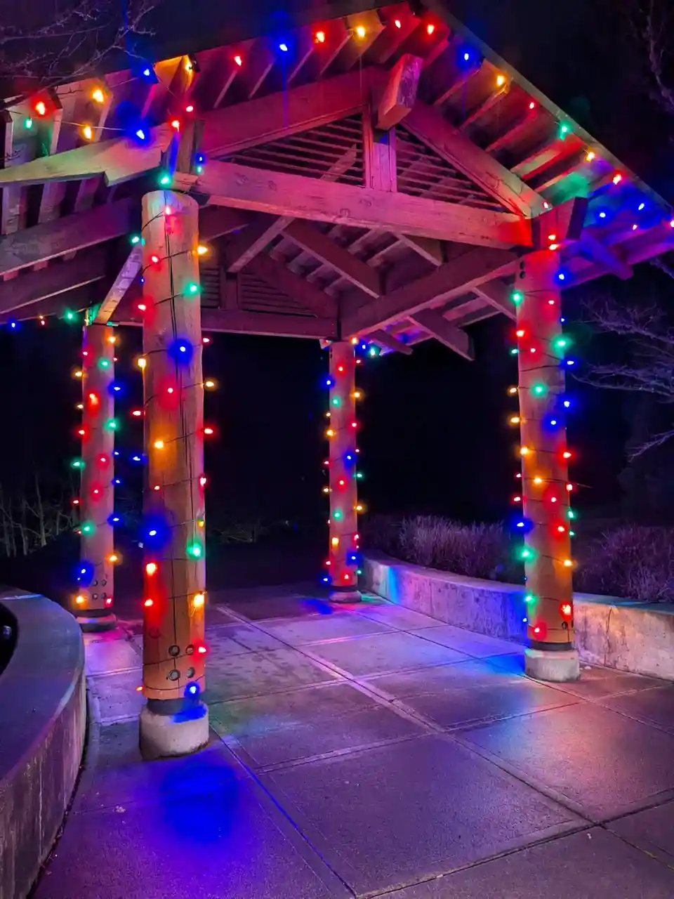 A nighttime photo of a wooden gazebo decorated with multicolored string lights wrapped around its pillars and along the roof…