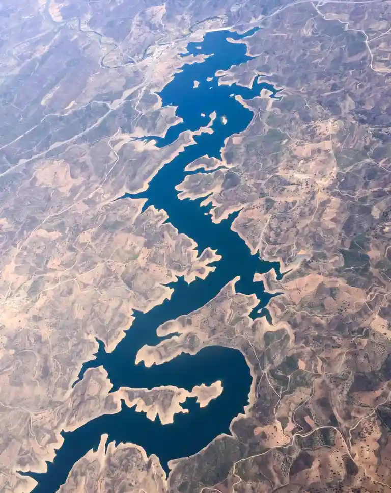 An aerial view shows a large, dark blue lake winding through arid, brownish terrain with visible roads and sparse…