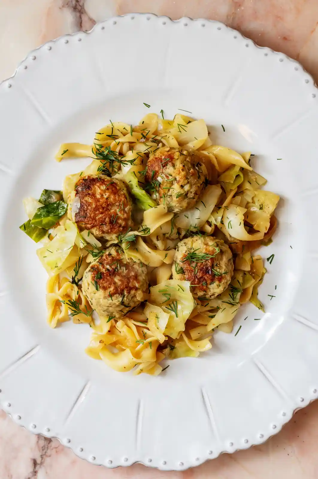 A top-down photo of a white plate with four browned meatballs, wide pasta noodles, and sautéed cabbage, garnished with…