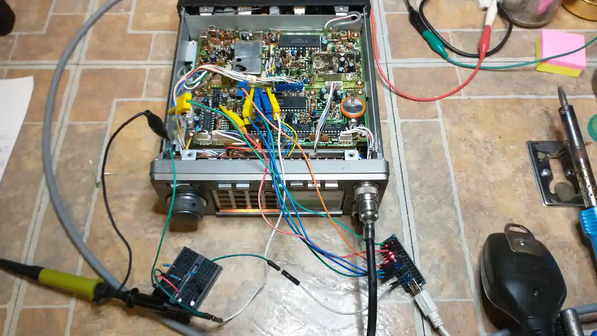 An open electronic device, likely a radio or audio unit, sits on a tiled floor surrounded by wires, a soldering iron, a…