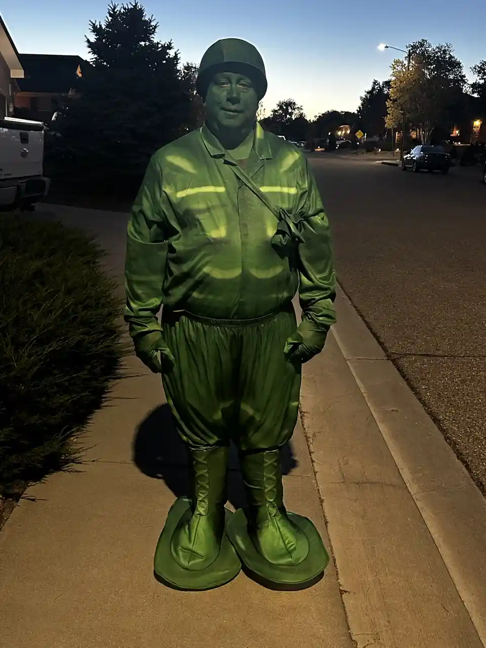 A person stands on a sidewalk at dusk wearing a full-body green military-style costume with helmet, gloves, and oversized…