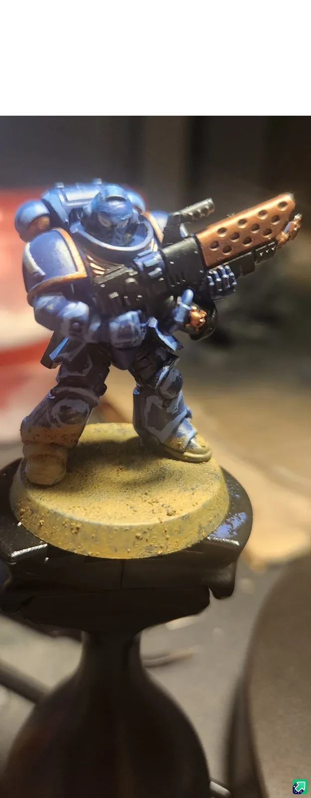 A close-up photo of a painted miniature of a Space Marine in blue and gold armor, holding a large, perforated weapon…