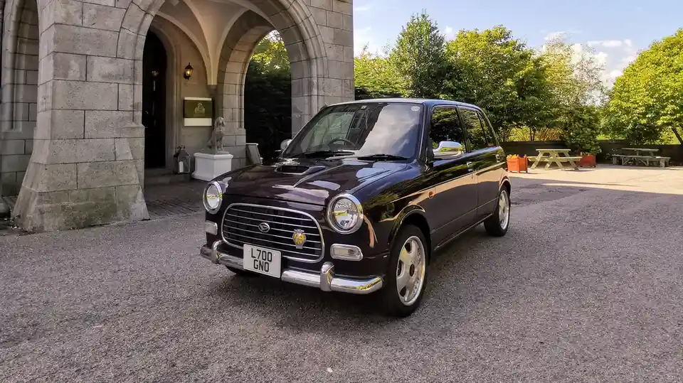 A glossy black, retro-styled compact car with chrome accents and a vintage grille is parked on gravel in front of a grand…