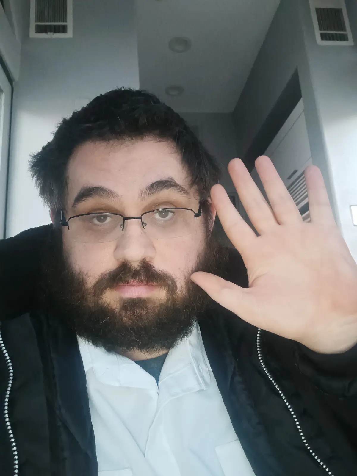 A man with a beard and glasses holds up his left hand with all five fingers extended toward the camera, wearing a black…