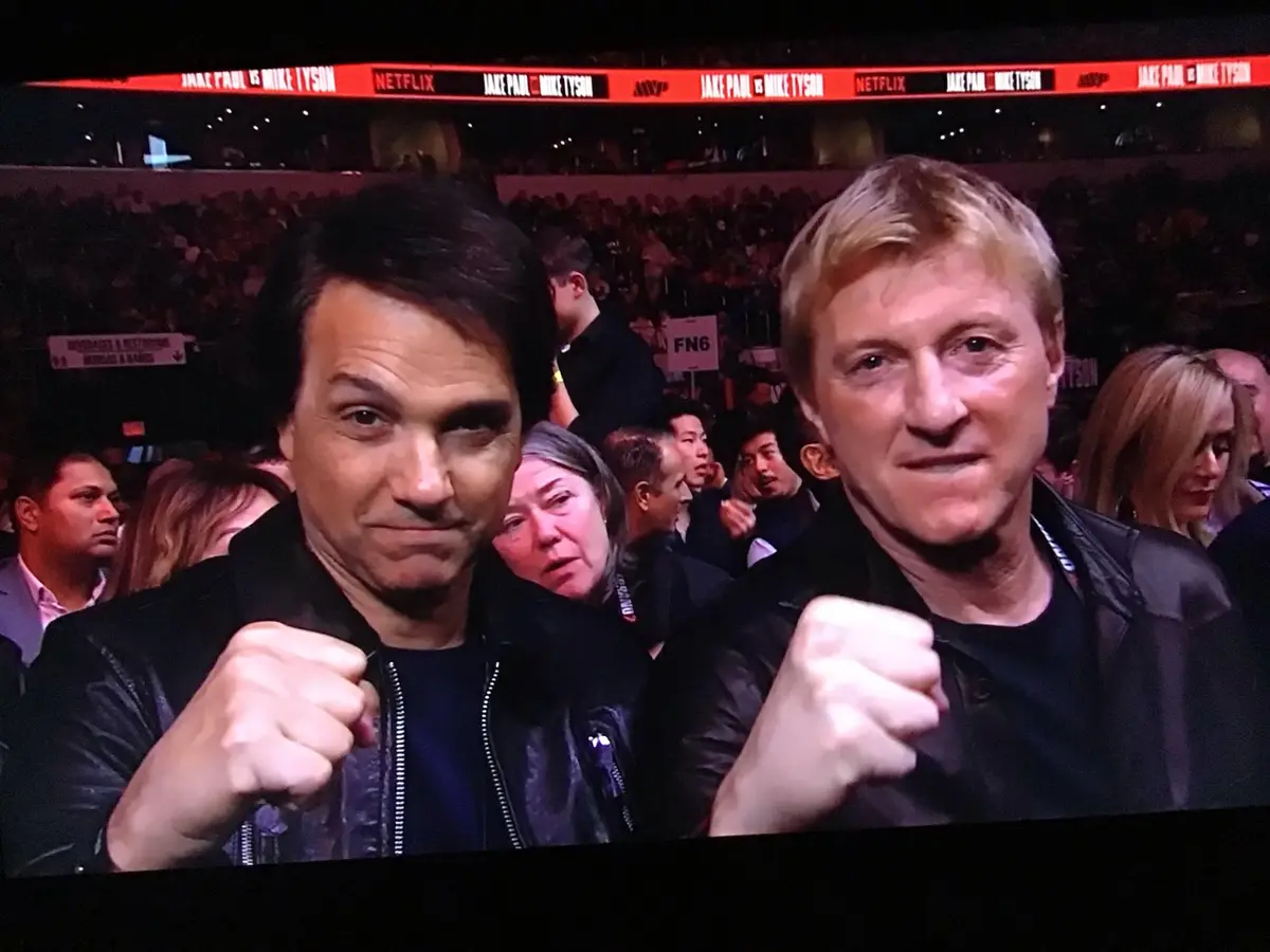 Two men, one with dark hair and one with blond hair, both wearing black jackets and holding fists up in a fighting pose, are…