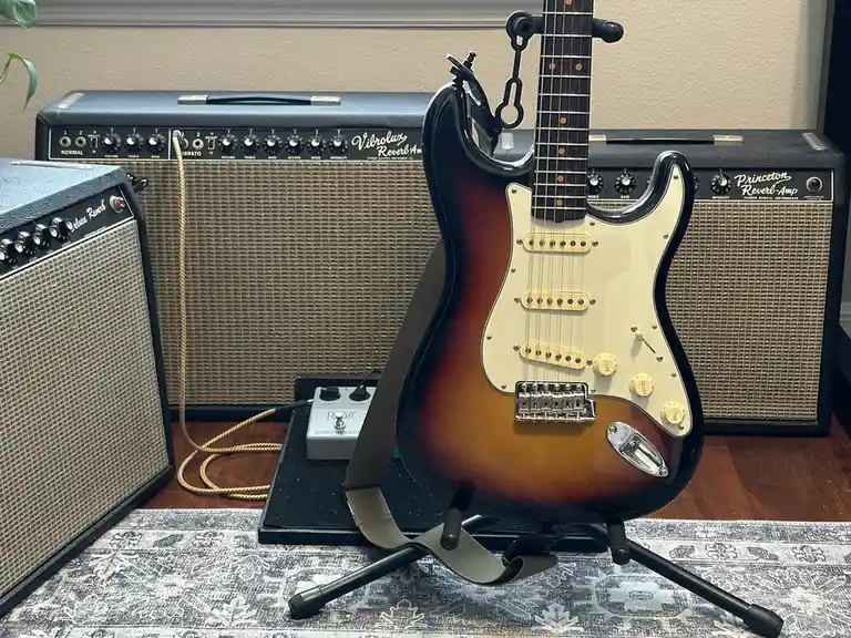 A sunburst-finish electric guitar stands on a stand in front of three vintage Fender amplifiers, with a Ross Compressor…
