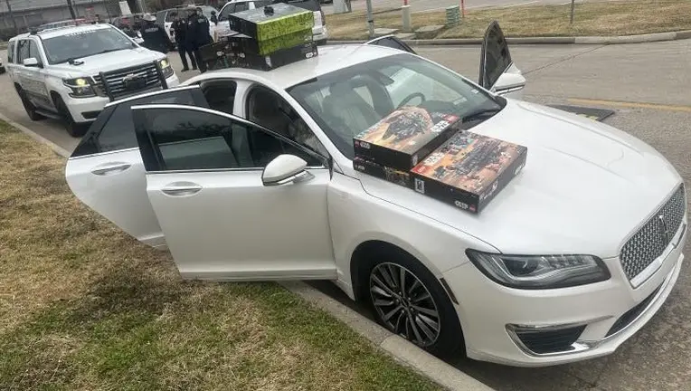 A white Lincoln sedan has its doors open and two large model kit boxes stacked on its hood. In the background, police…
