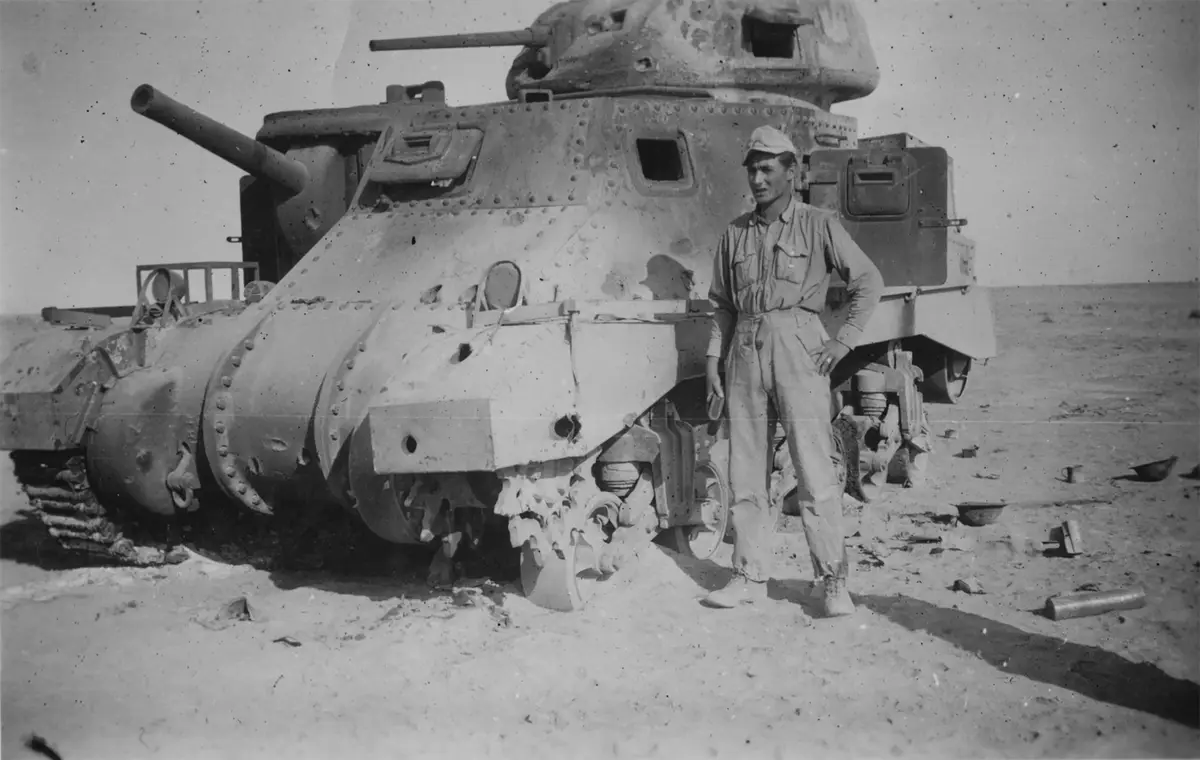 A black-and-white historical photo shows a soldier standing confidently beside a heavily damaged, battle-scarred tank in a…