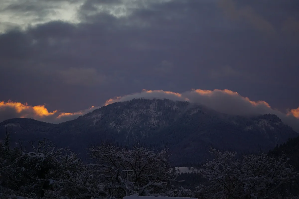 A snowy mountain landscape at dusk or dawn, with dark clouds and orange-tinged clouds on the horizon. Snow-covered trees and…