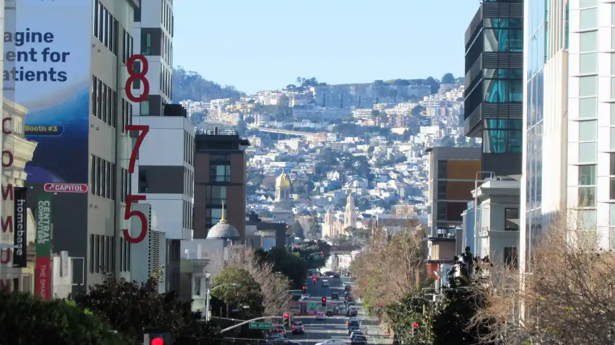 A daytime cityscape photo of a steep street in San Francisco, flanked by modern buildings with large red numbers '875'…